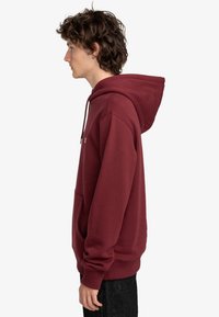Bordó mikina vyrobená z bavlny, obsahující klokaní kapsu, žebrované manžety a lem, s kapucí na stahovací šňůrku. Boční pohled, jednoduchý design.
