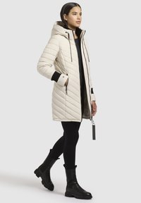 Lys beige quiltet jakke med hætte, med sorte detaljer på manchetter, lynlåslommer og en længere silhuet.