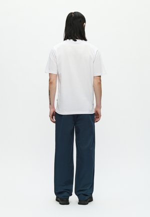 Camiseta blanca de algodón de manga corta vista desde atrás. Combinada con pantalones sueltos azul marino y zapatos negros. Diseño minimalista.