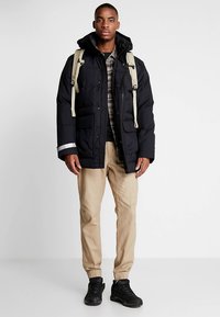 Zwarte donzen jas met afneembare capuchon, gecombineerd met beige joggerbroeken en zwarte sneakers. De jas heeft zakken aan de voorkant en reflecterende accenten.