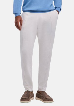 Mann trägt weiße Slim-Fit-Hosen, blauen Pullover und braune Wildleder-Slipper, steht mit einer Hand in der Tasche.