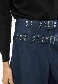 Tumesinised teksapüksid on varustatud laia vöökohaga, millel on metallist silmused ja hõbedane pandlaga. Kangas on sileda tekstuuriga ja hästi vormitud istuvusega.