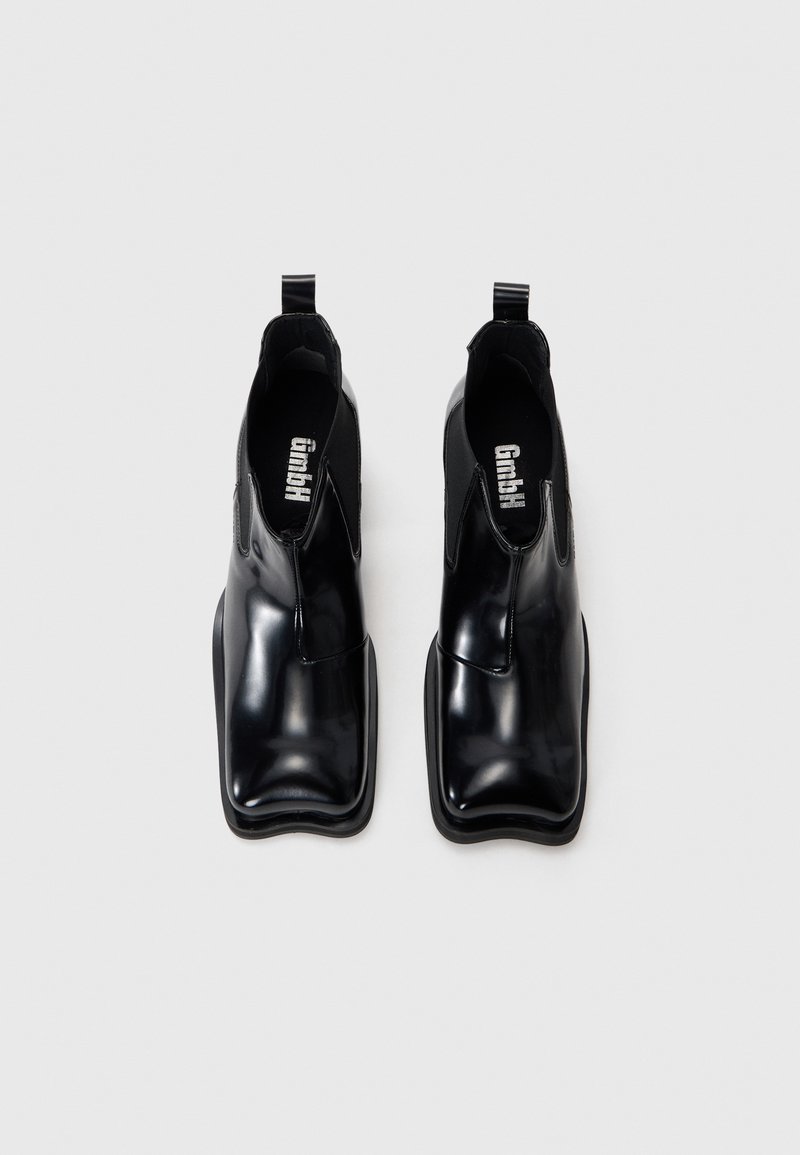 Bottines noires à talon avec finition brillante en vernis, panneaux latéraux élastiques, languettes de tirage et design à bout carré. Semelle en caoutchouc robuste avec un minimum de texture.