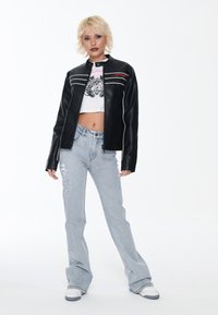 Giacca in ecopelle nera con strisce bianche e dettagli rossi, abbinata a una maglietta grafica corta e a jeans in denim chiaro strappati.