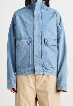 Person iført lyseblå denimjakke med høj krave og forlommer med klap, kombineret med beige bukser mod en ensfarvet baggrund.