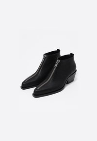 Botas de cuero negro hasta el tobillo con un diseño de punta puntiaguda, que presentan una prominente cremallera plateada en la parte delantera y un tacón ancho.