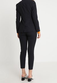 Skräddarsydd svart kavaj med en åtsittande silhuett och orange manschett, matchad med smala svarta byxor. Har stilrena, högklackade vita skor.