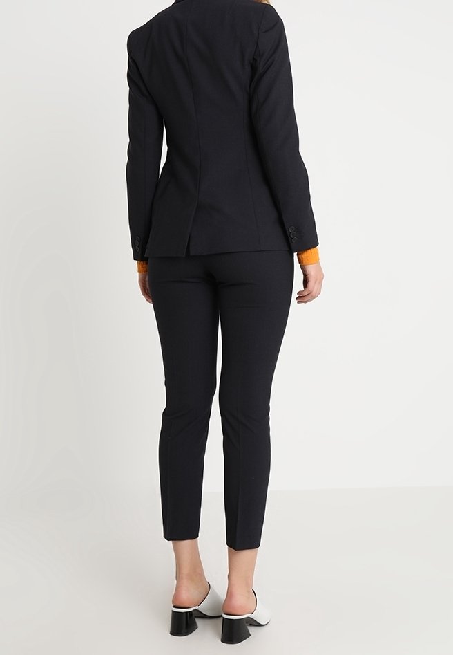 Skräddarsydd svart kavaj med en åtsittande silhuett och orange manschett, matchad med smala svarta byxor. Har stilrena, högklackade vita skor.