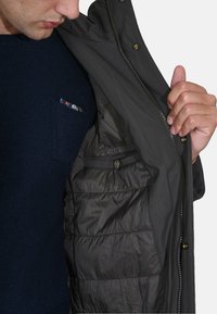 Giacca grigio scuro con tessuto esterno liscio, internamente foderata, chiusura con zip e tasche interne. Indossata aperta sopra un maglione blu navy.