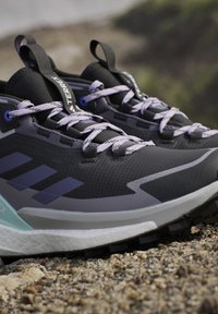 Gros plan sur des chaussures de trail running Adidas Terrex noires et grises avec des lacets violets sur un sol extérieur rocheux.