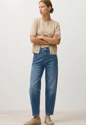 Woman stands with arms crossed, wearing a beige short-sleeve cardigan, light brown T-shirt, blue jeans, and beige sneakers.