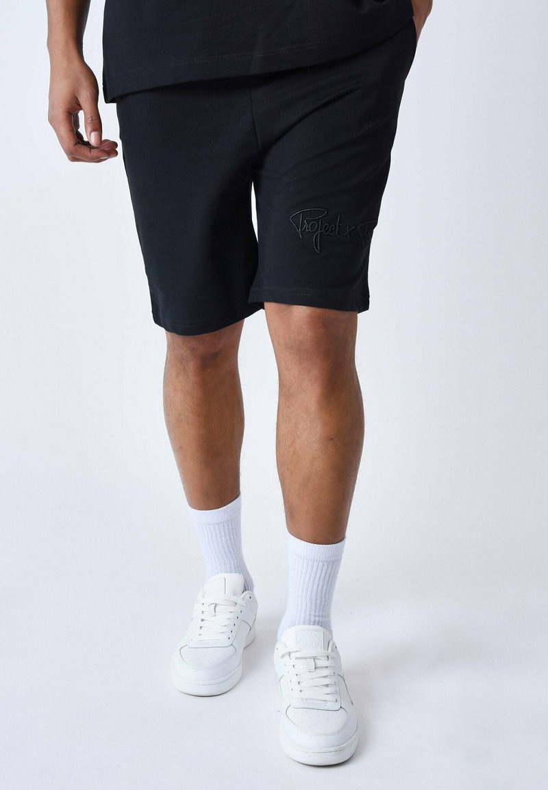 Shorts en coton noirs au design ajusté, avec une marque cousue sur la jambe gauche. Associés à des chaussettes blanches montantes et des baskets blanches.