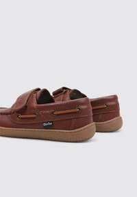 Zapatos de cuero marrón con punta redonda, una correa de velcro y cordones decorativos. Suela de goma marrón texturizada con un perfil bajo.