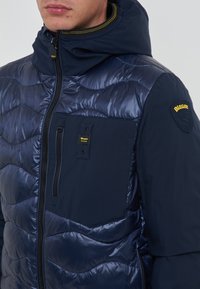 Marineblauwe gewatteerde jas met een glanzende afwerking, voorzien van een capuchon, een rits aan de voorkant en een borstzak met gele accenten en logo.