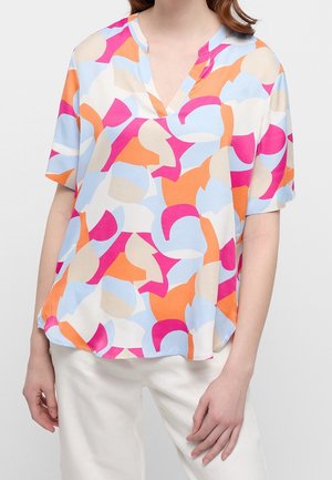 Kobieta nosząca luźną bluzkę z krótkim rękawem z wielobarwnym abstrakcyjnym wzorem w kolorze różowym, pomarańczowym, niebieskim i beżowym, w połączeniu z białymi spodniami.