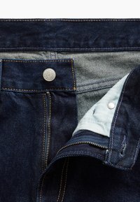 Σκούρο μπλε τζιν παντελόνι με ανοιχτό φερμουάρ και κουμπί, που δείχνει το ύφασμα της εσωτερικής τσέπης και λεπτομερείς πορτοκαλί ραφές.