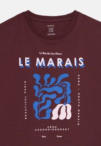 Tmavě červené tričko s modro-bílým abstraktním květinovým vzorem a textem "Le Marché Aux Fleurs LE MARAIS" a odkazy na pařížské čtvrti.