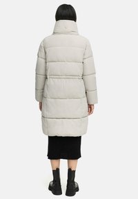 Abrigo acolchado beige con cuello alto, con secciones acolchadas horizontales y rematado con un detalle de botón en el cuello. Falda negra de canalé y botas.