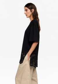 Must mustriga särk, millel on laineline pitsäär, lõdva istuvuse ja lühikeste varrukatega. Kombineeritud heledate laia säärtega pükstega.