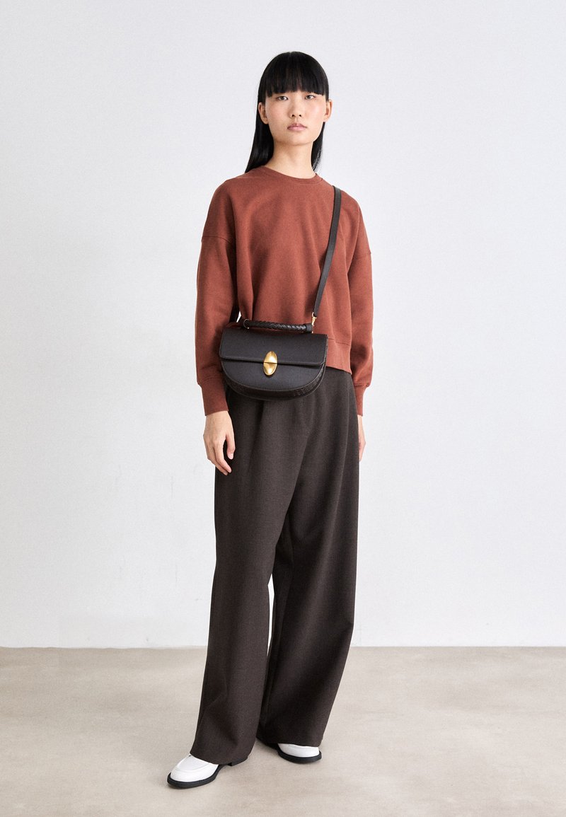 Femme avec de longs cheveux noirs portant un pull rouille, un pantalon large sombre, des chaussures blanches et un sac bandoulière noir avec un fermoir doré, se tenant à l'intérieur.