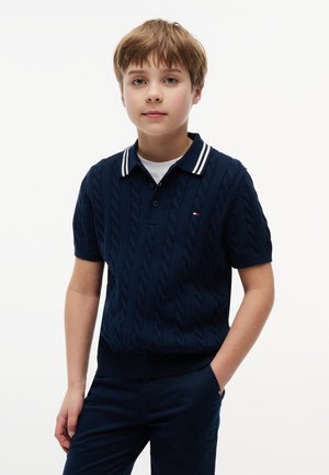 Αγόρι με ανοιχτό καστανά μαλλιά, φορώντας μπλε σκούρο πλεκτό πόλο με κοντά μανίκια και ταιριαστό παντελόνι, στέκεται με το ένα χέρι στην τσέπη.