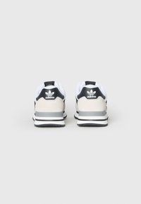 Sporta apavi ar baltu un pelēku augšu, melnām detaļām un teksturētu zolīti. Papēdī ir redzams logo, un vidējā zole ir ar svītrām.