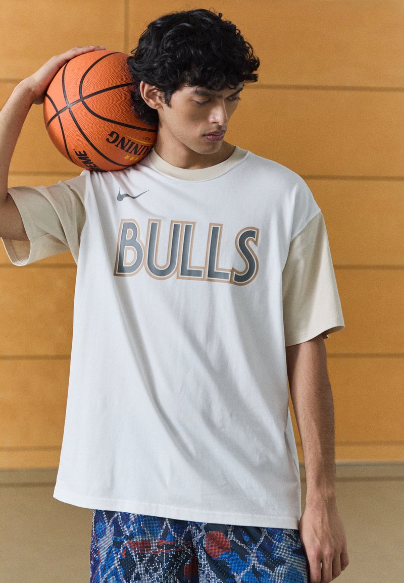 Overdimensioneret hvid og beige t-shirt med "BULLS" tekst, parret med mønstrede blå shorts. Personen holder en basketball på deres skulder.