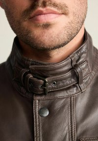 Braune Lederjacke mit hohem Kragen und Schnalle, strukturierter Oberfläche und einem runden Metalldetail nahe der vorderen Naht.