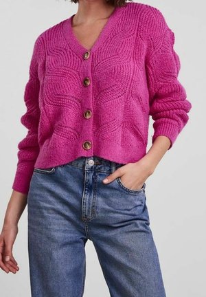 Femme portant un cardigan rose texturé boutonné à manches longues et un jean bleu taille haute, une main dans la poche, sur un fond uni.
