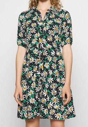 Mujer con vestido negro de manga corta con estampado floral de margaritas blancas y amarillas, cierre de botones al frente y cuello con solapa, de pie contra un fondo liso.