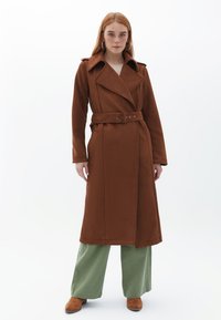 Brązowy płaszcz trench z szerokim kołnierzem i długimi rękawami, z dwoma bocznymi kieszeniami i miękką teksturą materiału. Model nosi oliwkowe spodnie.