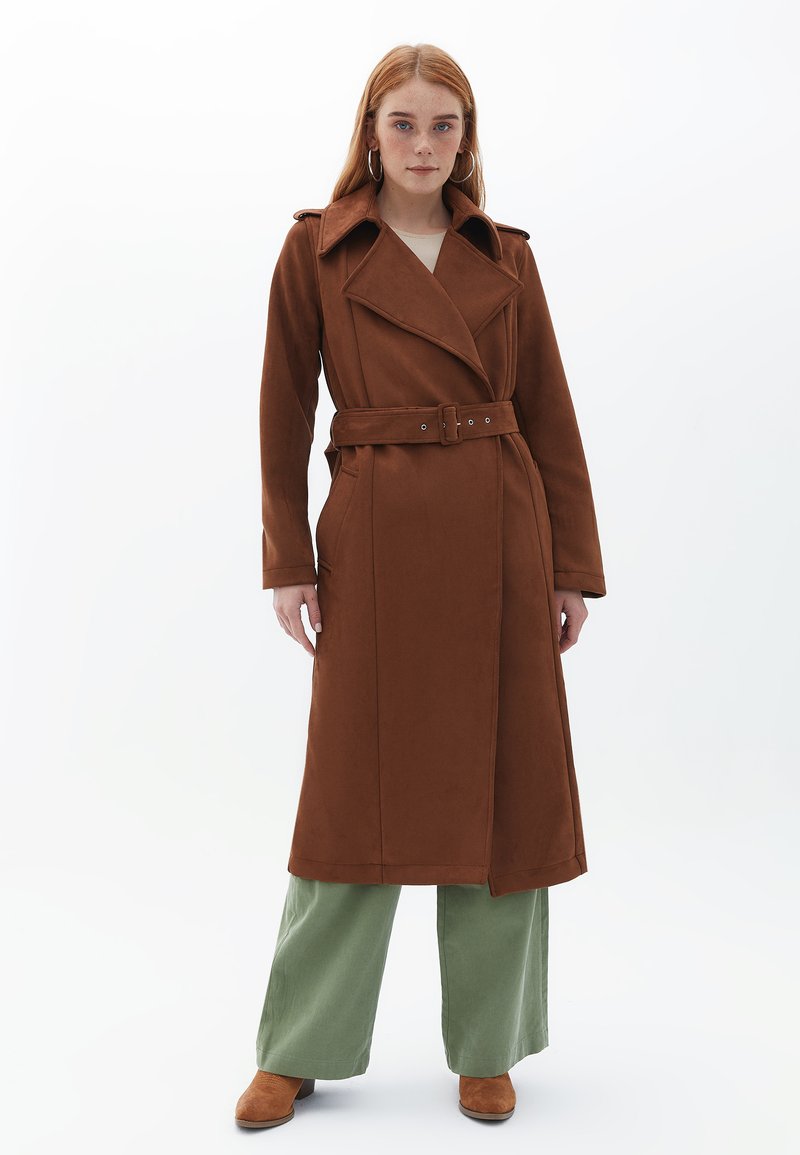 Gabardina marrón con cinturón, cuello ancho y mangas largas, que cuenta con dos bolsillos laterales y una textura de tejido suave. El modelo lleva pantalones de color verde oliva.