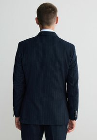 Granatowa marynarka w prążki o dopasowanym kroju, gładkiej fakturze i subtelnym połysku. Posiada pojedynczy rozcięcie i akcenty przy mankietach z czterema guzikami.