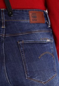 Gros plan sur la poche arrière d'un jean en denim bleu avec des coutures orange, un patch de marque en cuir, et un pull rouge visible à la taille.