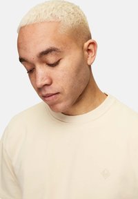 Hellbeige Crewneck-Sweatshirt aus weichem Material, mit einem dezenten, geprägten Logo auf der Brust und einem strukturierten, gerippten Kragen.