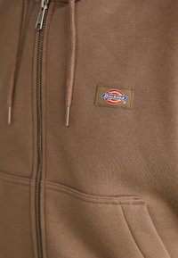 Pruun tõmblukuhoodie, mis on valmistatud pehmest materjalist, esiosas on tasku ja vasakul küljel Dickiesi logo paigaldus. Õrnad tikandid on nähtavad.