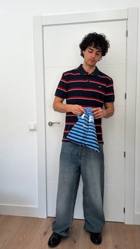 Jauns vīrietis svītrainā polo krekla un brīvās biksēs stāv pie baltām durvīm, turēdams salocītu zilu un melnu svītrainu audumu.