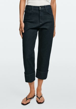 Kobieta w czarnych podwiniętych spodniami typu cropped jeans, białym topie i czarnych klapkach stojąca na tle jednolitego tła.