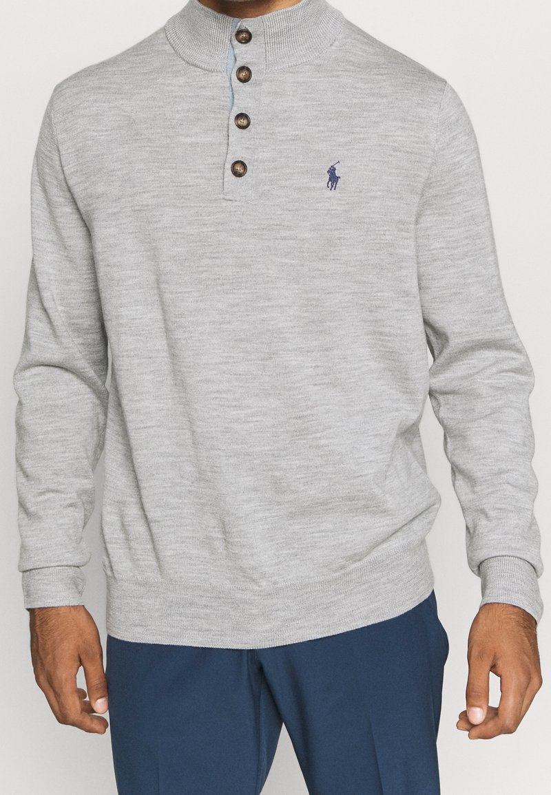 Homme portant un pull henley à manches longues gris clair avec quatre boutons marron et un pantalon bleu marine, se tenant devant un fond uni.