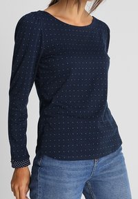 Marineblauwe longsleeve top met kleine blauwe stippen, ronde halslijn en gestructureerde mouwen. Gepaard met lichtblauwe denim jeans.