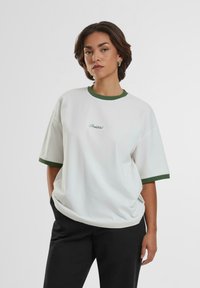 Camiseta blanca de gran tamaño con cuello redondo acanalado verde, mangas cortas y texto bordado en verde en la parte frontal. Combinada con pantalones negros.
