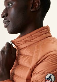 Giubbotto a gilet arancione con texture trapuntata, colletto alto e zip. Presenta una patch con logo sulla spalla sinistra. Superficie del tessuto liscia e lucida.