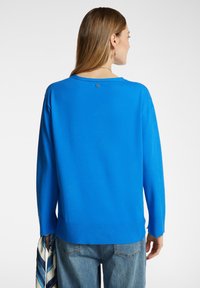 Blauwe gebreide trui met lange mouwen en een ronde halslijn. Heeft subtiele stikdetails aan de zoom en een klein logo op de achterkant.