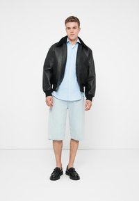 Fekete bőr bombázó dzseki galléros nyakkal, világoskék inggel és világos farmer rövidnadrággal viselve, fekete cipőkkel kiegészítve.