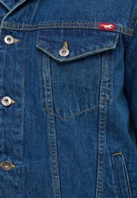 Niebieska kurtka dżinsowa o klasycznym fasonie, z dwiema kieszeniami na piersi, srebrnymi guzikami i kontrastowymi przeszyciami. Czerwona metka z tkaniny na lewej kieszeni.