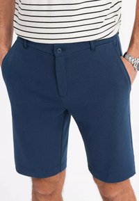 Marineblaue Shorts aus strukturiertem Stoff, mit einem vorderen Knopfverschluss, Seitentaschen und einem geraden Saum. Kombiniert mit einem gestreiften Oberteil.
