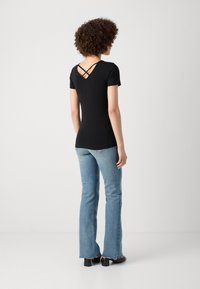 Femme aux cheveux bouclés portant un haut noir à manches courtes avec des bretelles croisées dans le dos et un jean bleu clair, debout sur un fond blanc.