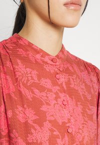 Blusa rossa con motivo floreale, dalla finitura testurizzata, con colletto alla mandarino e bottoni, evidenziando delicati disegni di rose in rosa.