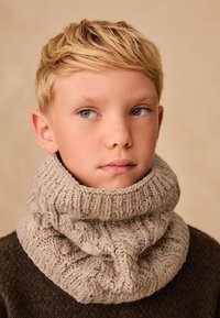 Bézs kötött nyakmelegítő texturált kötélszerű mintával és bordázott felső szegéllyel, sötét pulóver fölött viselve. Sima felület, kényelmes megjelenés.