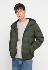 Giovane uomo con una giacca a vento verde scuro aperta, un maglione grigio chiaro e jeans blu, in piedi contro uno sfondo bianco semplice.